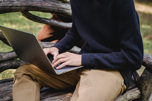 Person sitting on a wooden bench outdoors typing on a laptop